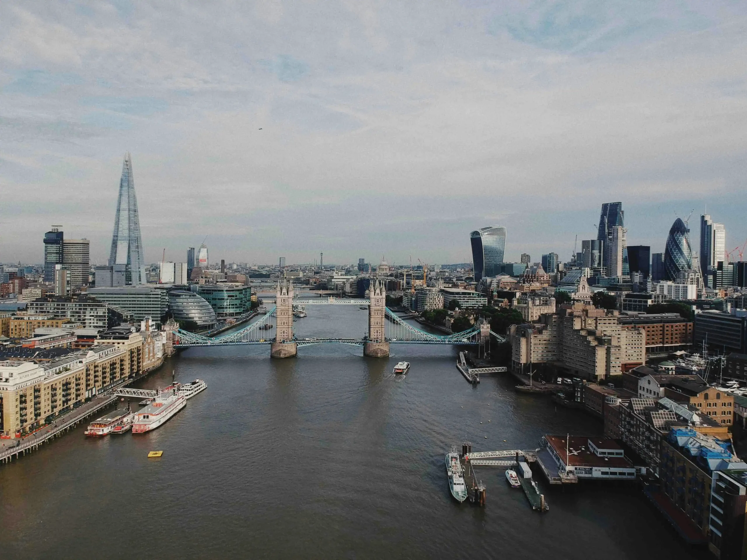 Wide aerial drone photo looking down the River Thames toward Tower Bridge and the central London skyline.