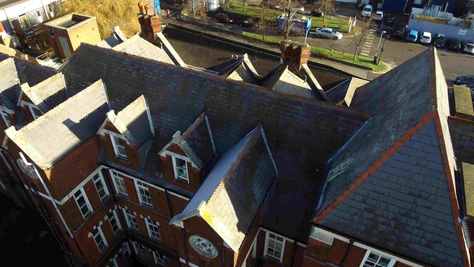 High-angle aerial drone view inspecting the pitched slate tile roof, gables, and chimneys of a historic red brick building.