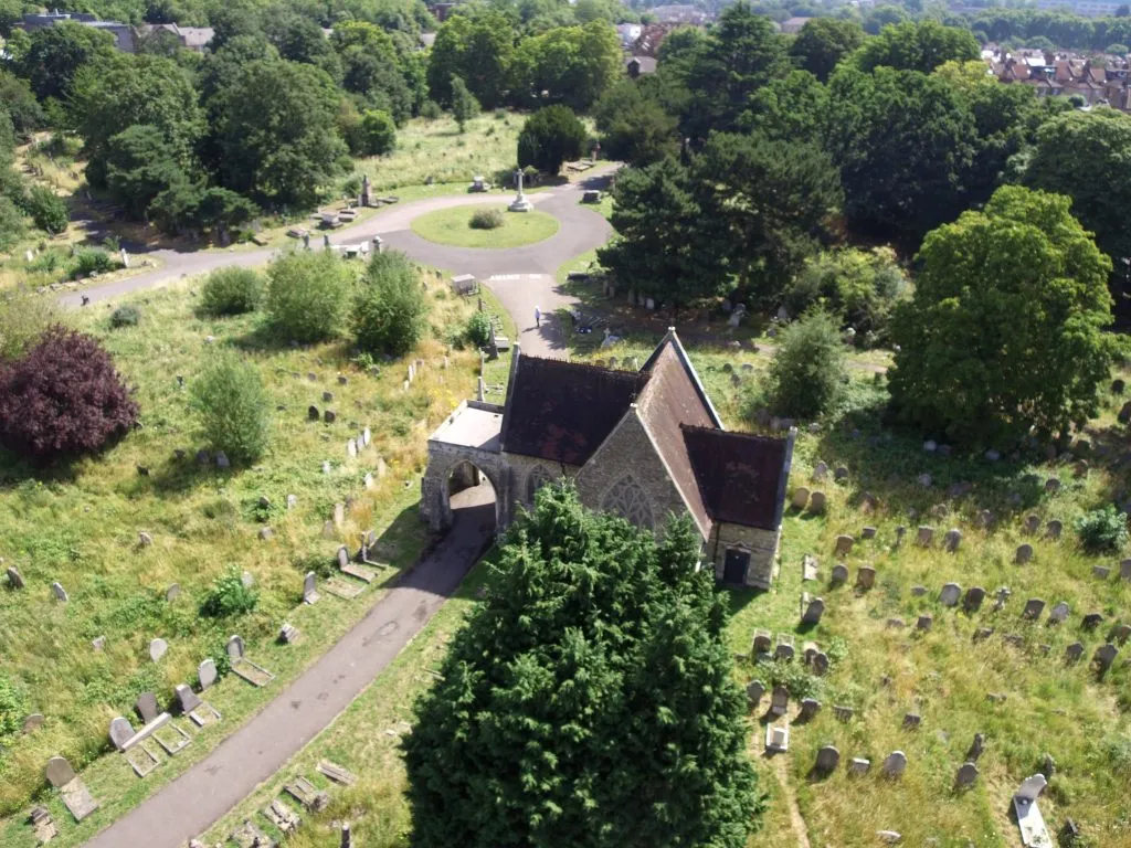 High-angle aerial drone view of a stone chapel building surrounded by headstones and trees in a cemetery.