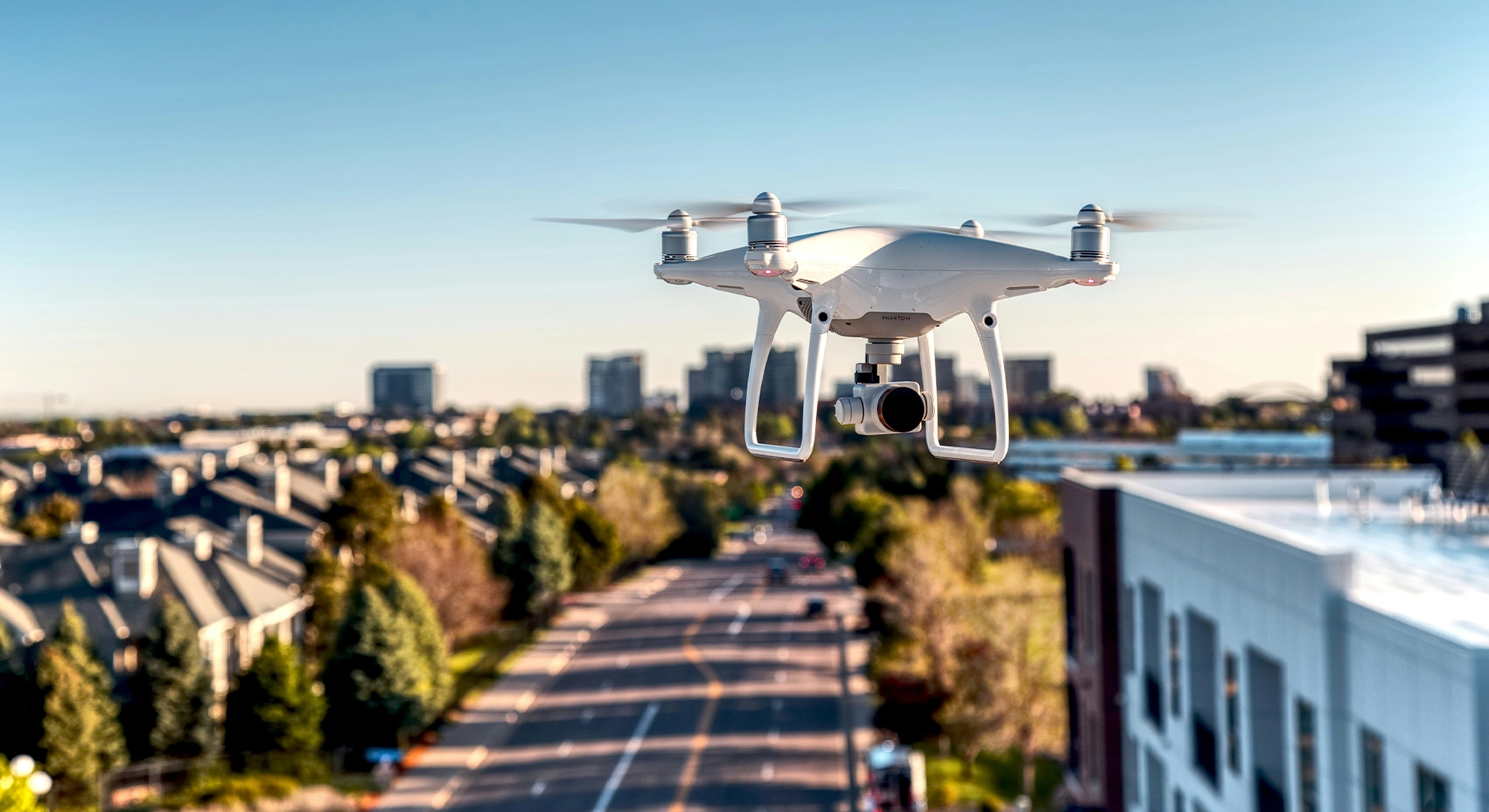 Close-up shot of a white quadcopter drone hovering in flight with its camera trained forward. The blurred background reveals a sunny residential neighbourhood, trees, and a road under a clear blue sky.