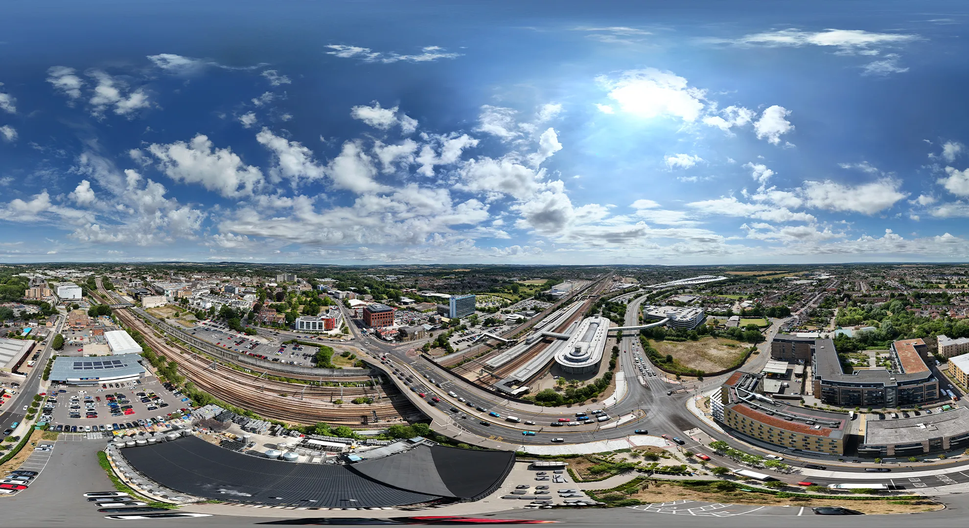 Ultra-wide 360-degree aerial drone panorama of a city showing train tracks, highways, buildings, and a bright cloudy sky.