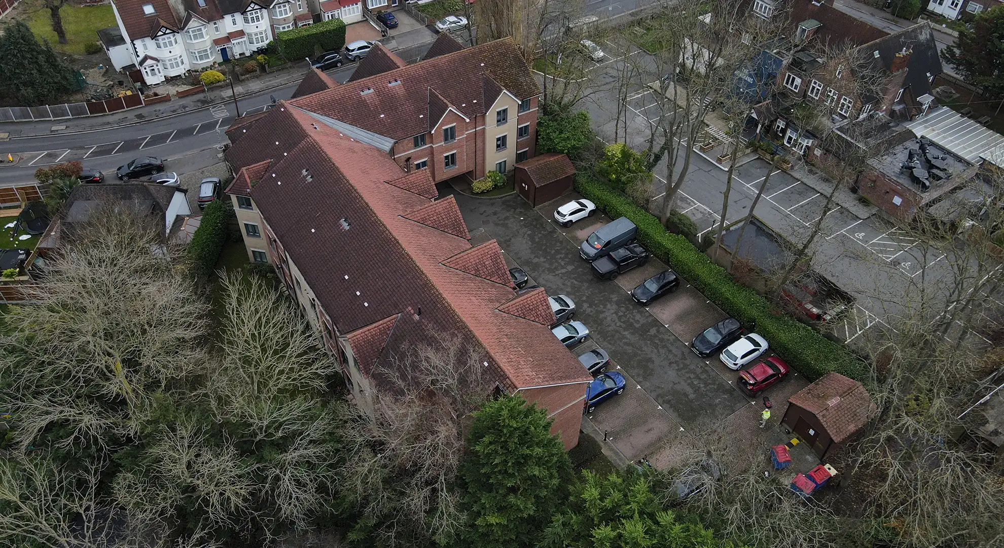 Overhead drone view showing the roof architecture and layout of a multi-story brick apartment complex with red tiled gables. Below, several cars are parked in an internal courtyard, surrounded by surrounding trees and nearby roads.