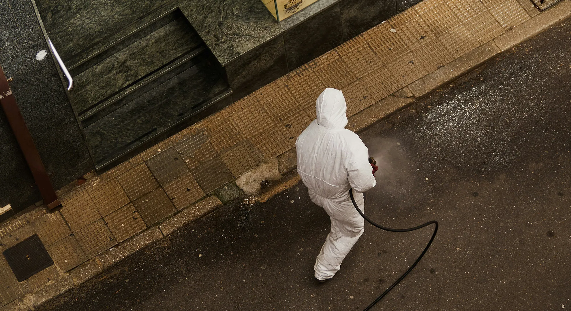 Aerial view of a worker in a full white hazmat suit spraying disinfectant along a city street outside a shop.