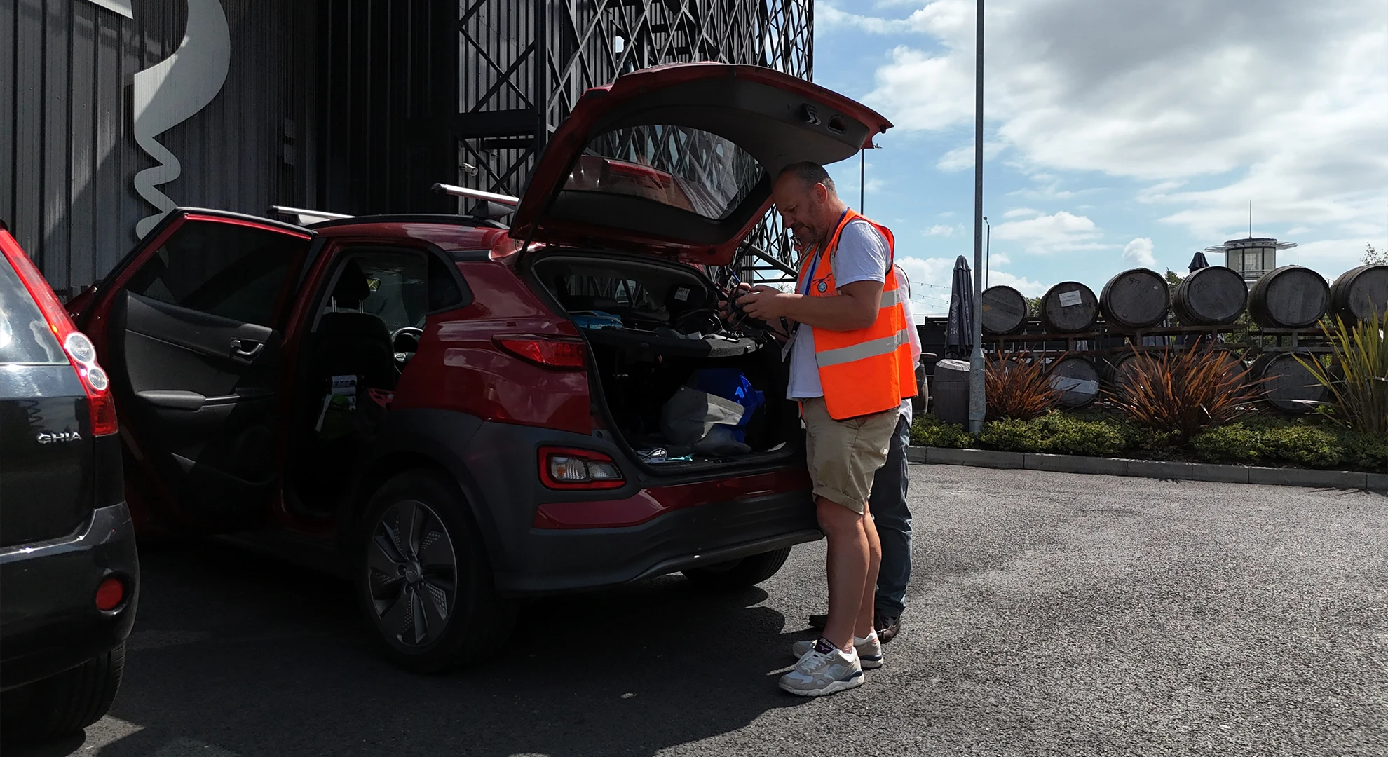 Helidrone Pilot Bob McGowan, wearing an orange safety vest, looks into the open trunk of a red SUV parked near a building and barrels