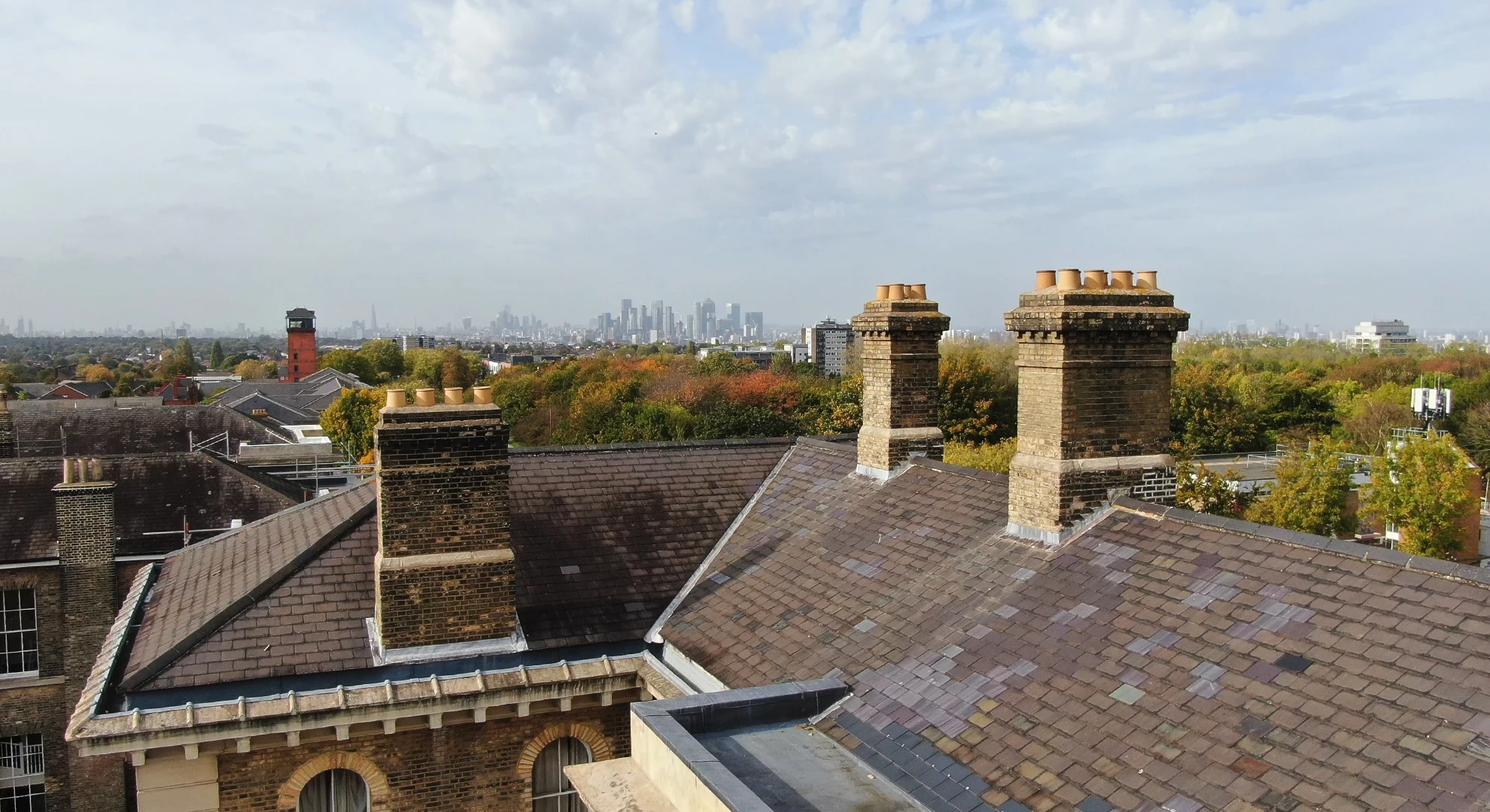 Drone view of brick chimneys and slate roofs overlooking autumn trees with a distant London skyline