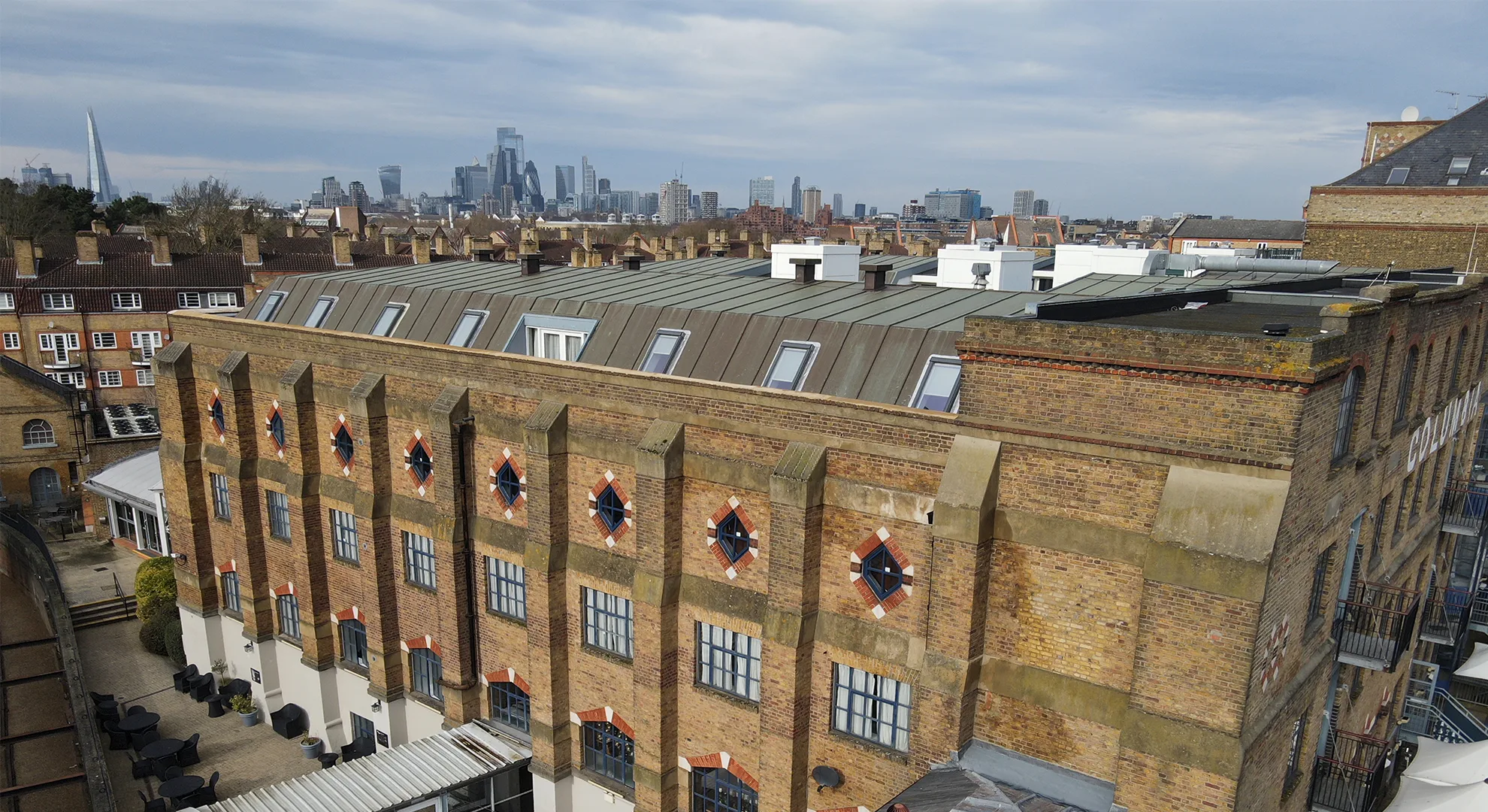 Drone aerial photo of a large brick building with diamond-shaped windows and skylights, with the London skyline in the background