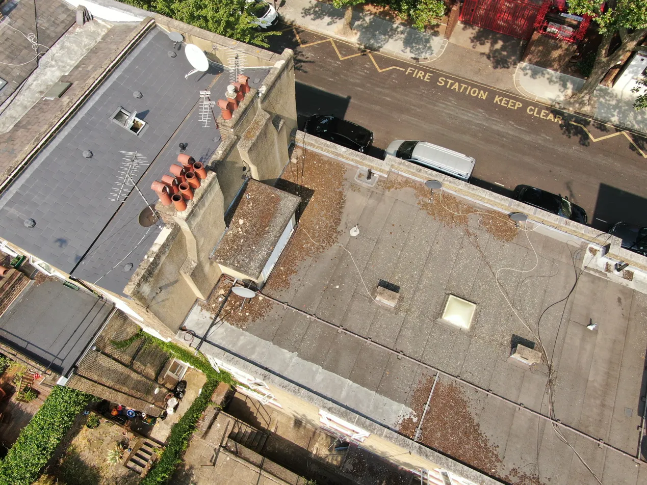 Drone aerial view of a flat rooftop with multiple chimney pots and satellite dishes, overlooking a city street