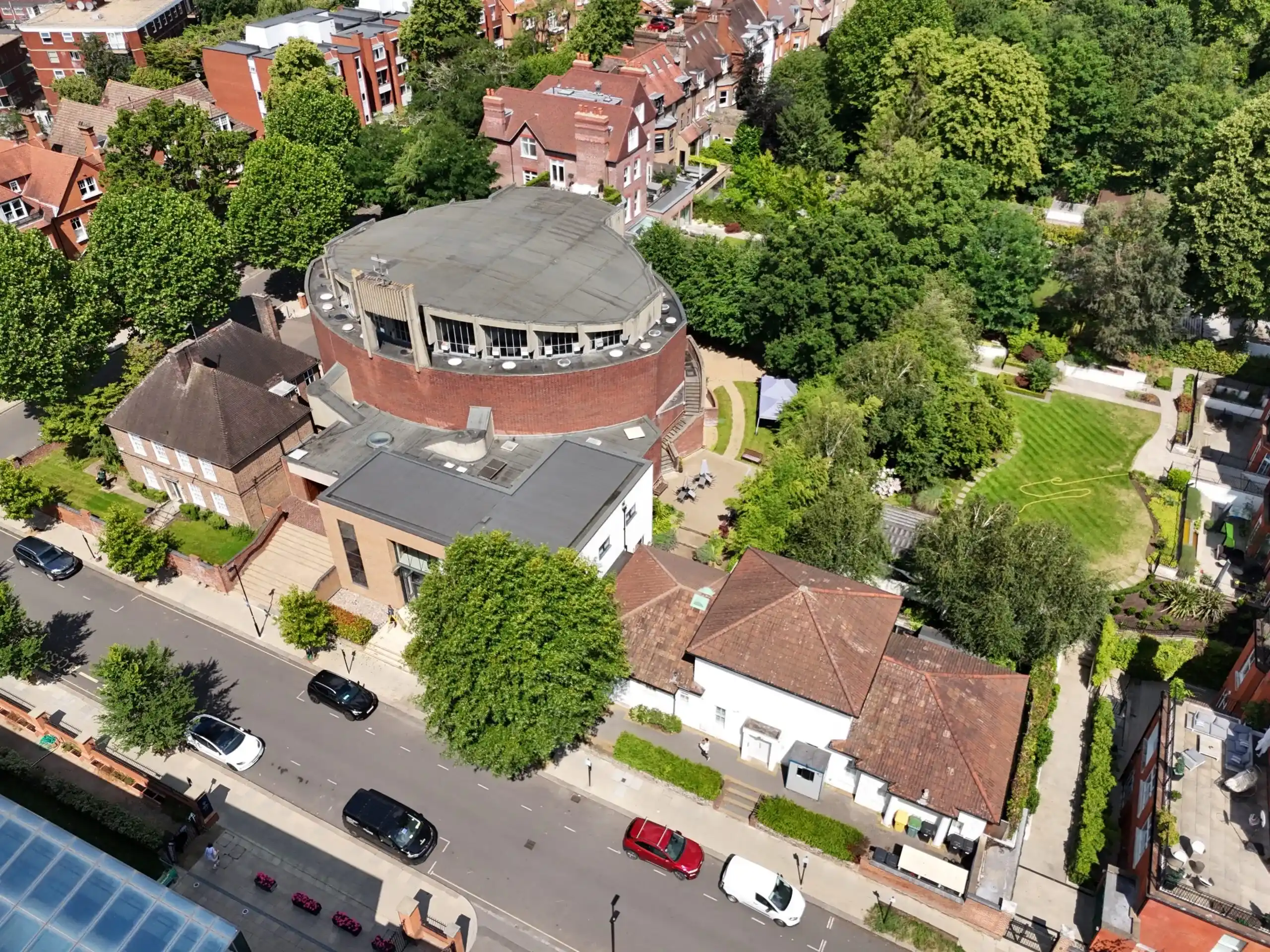 High-angle aerial drone view of a circular brick building complex showing roof structure, surrounding trees, and adjacent street.