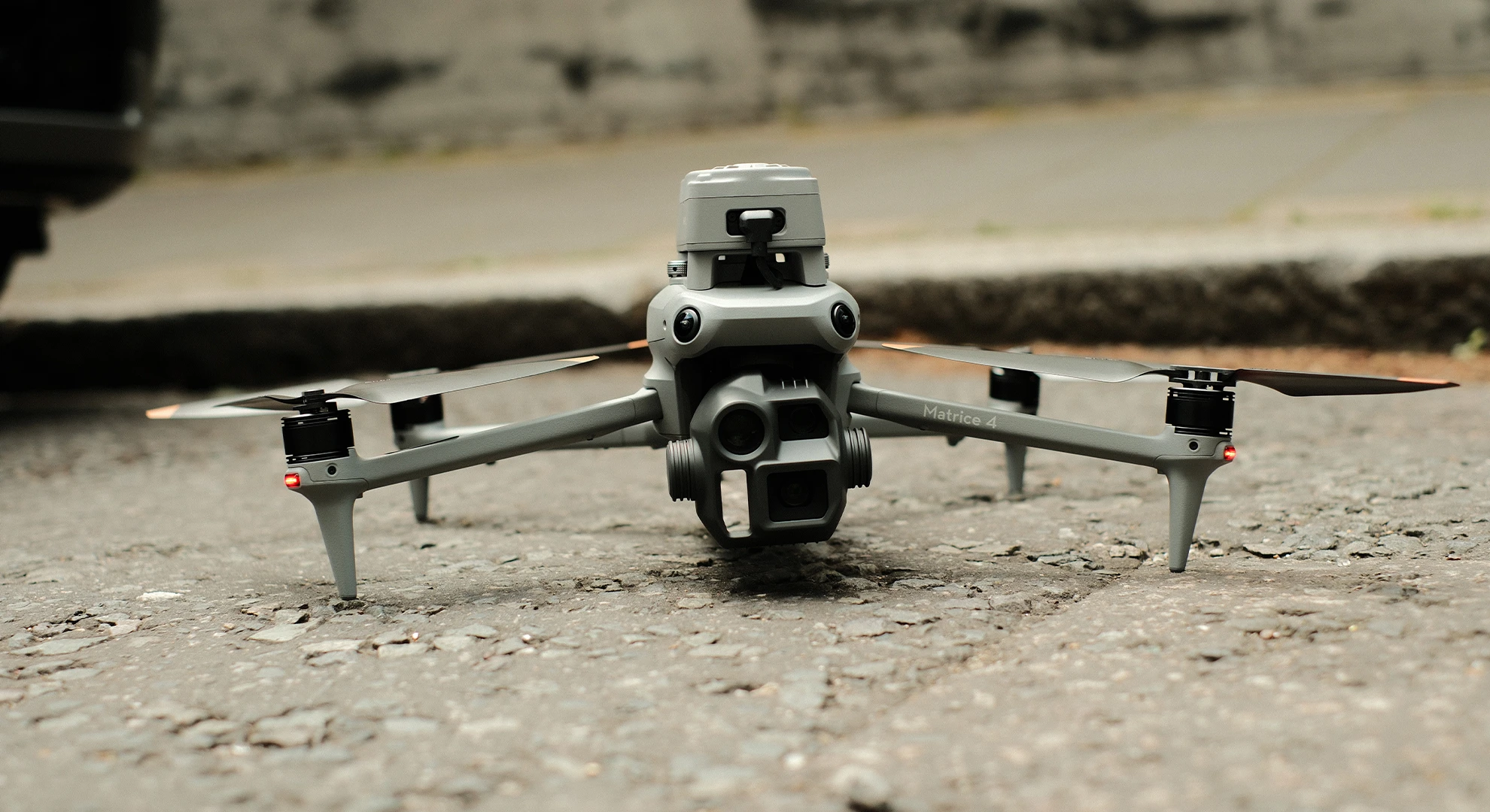 Front view of HeliDrone's grey DJI Matrice 4 drone on asphalt, showing its multi-lens camera gimbal and sensors