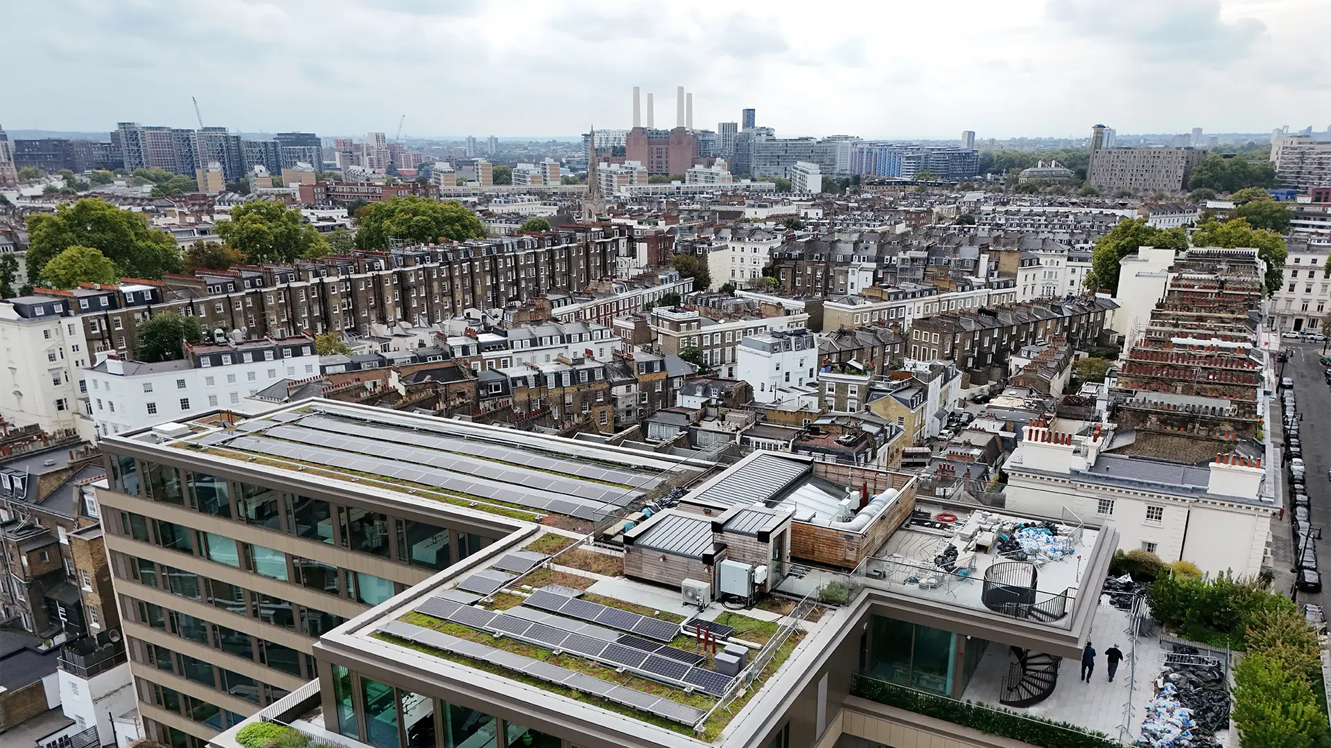 Elevated aerial drone view of a modern building rooftop with solar panels and a green roof, set against an urban city backdrop.